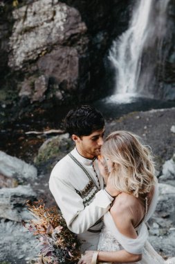 Ses efekti, seçici odak: İnanılmaz derecede aşık gelinlerin kucaklaşması, öpüşmesi ve poz vermesi inanılmaz kayalık arka planda büyük bir şelale ile birlikte.