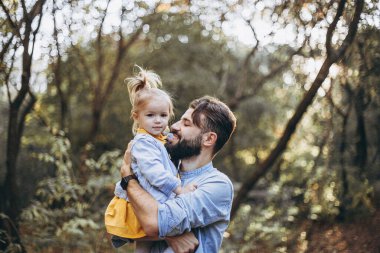Genç, şık sakallı bir baba gün batımında küçük kızıyla birlikte doğada yürüyor. Baba ve çocuğun aile fotoğrafları. Babalar günün kutlu olsun. seçici odak, gürültü etkisi