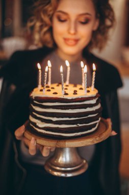 Güzel bir gülüşü olan çekici sarışın kız pasta tutuyor. Beklenmedik bir hediyeden mutlu bir doğum günü kızının duyguları: yanan mumlu ev yapımı pasta, seçici odaklanma, gürültü efekti