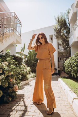 Turuncu bir pantolon ve güneş gözlüğü takan şık, genç ve çekici bir kadın yazın palmiye ağaçlarının ve parlak güneşin altındaki denizin arka planında fotoğraflandı.