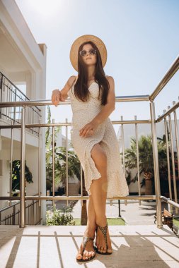 Hafif bir yaz elbisesi giyen şık, genç ve çekici bir kadın yazın arka planda palmiye ağaçları ve parlak güneşin altındaki deniz üzerinde fotoğraflandı.