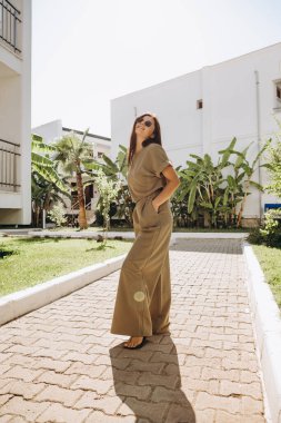 Tişört ve zeytin rengi pantolon giymiş şık ve çekici bir kadın yazın palmiye ağaçlarının arka planında ve parlak güneşli denizde fotoğraflandı.