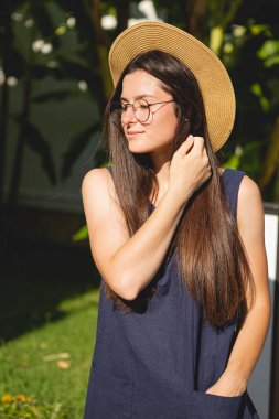 Şık, genç ve çekici bir kadın, mavi bir kısa günlük elbise ve şapka giyer ve yazın arka planda palmiye ağaçları ve parlak güneş altındaki deniz üzerinde fotoğraf çekilir.