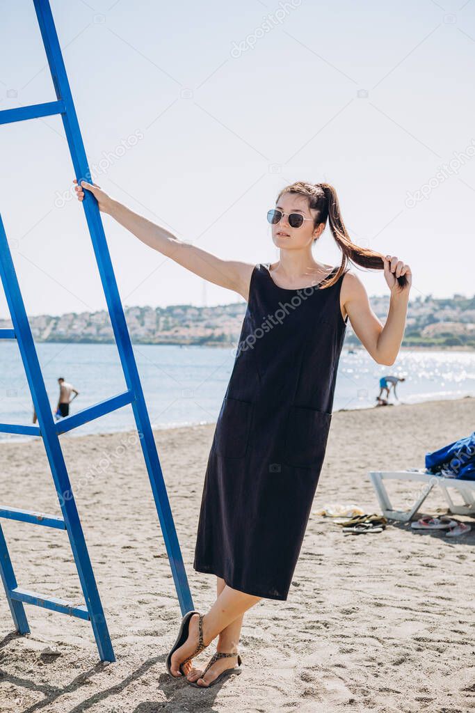 stylish young attractive woman dressed in wearing a blue short casual dress and hat photographed in the summer on a background of palm trees and the sea in the bright sun