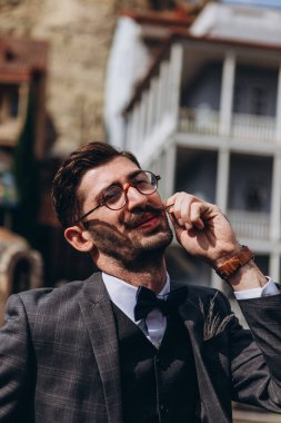 Tiflis 'teki eski şehrin arka planında fotoğraf için poz veren bıyıklı ve klasik smokinli şık genç bir Gürcü' nün portresi.
