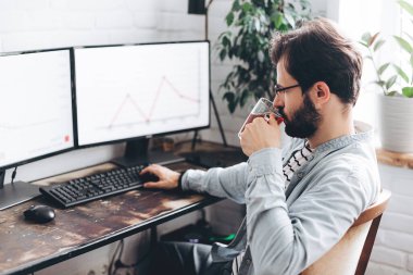 Gözlüklü genç işadamı bilgisayar monitörlerindeki grafikleri kontrol ediyor. Gözlüklü, konsantre olmuş milenyum adamı evde kendini izole etme üzerine çalışıyor.