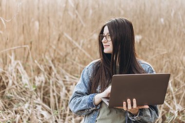 Genç bir kadın doğada dizüstü bilgisayarla çalışıyor. Açık hava doğa yolculuğu ve rahatlama. Serbest çalışma kavramı.