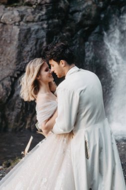 Ses efekti, seçici odak: İnanılmaz derecede aşık gelinlerin kucaklaşması, öpüşmesi ve poz vermesi inanılmaz kayalık arka planda büyük bir şelale ile birlikte.