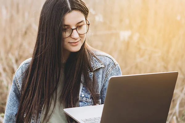 Happy hipster young woman working on laptop in field. Outdoors nature journey and relaxation. Freelance work concept.