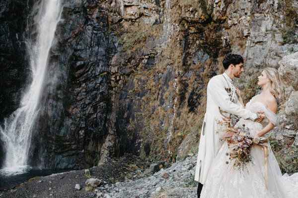 Noise effect, selective focus: incredibly enamored brides hugging, kissing and posing for a photo on the incredible rocky mountains background with a big waterfall