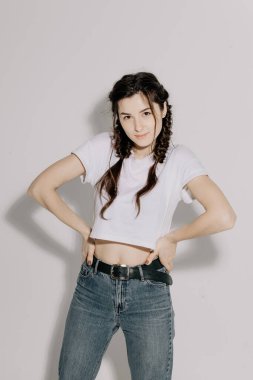 Beyaz tişört ve kot pantolon giymiş atölye ışığı önünde beyaz arka planda fotoğraf için poz veren at kuyruklu güzel bir kadın.