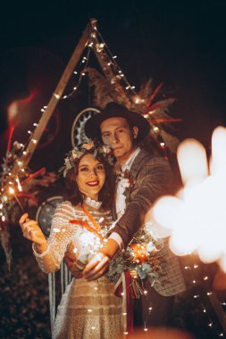 Akşam parktaki düğün fotoğrafında parlak ışıklar ve maytaplarla şık bir çift gelin.