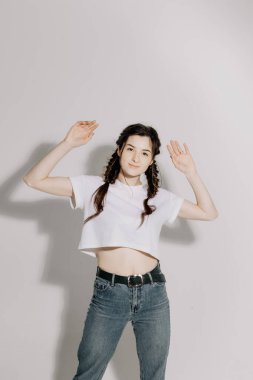 Beyaz tişört ve kot pantolon giymiş atölye ışığı önünde beyaz arka planda fotoğraf için poz veren at kuyruklu güzel bir kadın.