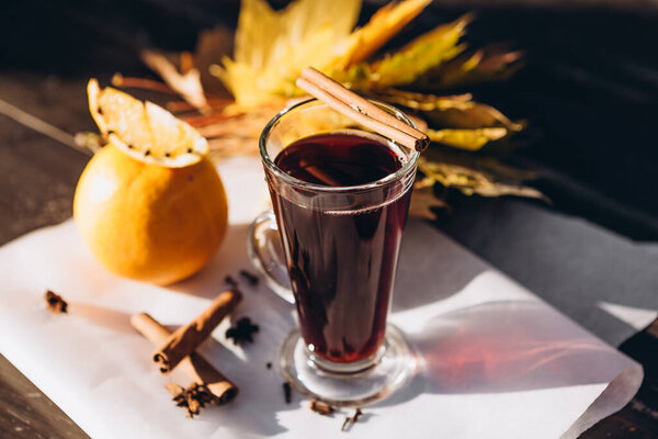 Transparent tall glass with red mulled wine boiled from grape wine on wooden background with autumn leaves, oranges and spices