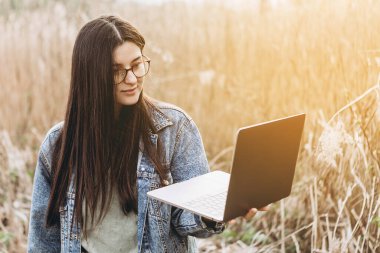 Genç bir kadın doğada dizüstü bilgisayarla çalışıyor. Açık hava doğa yolculuğu ve rahatlama. Serbest çalışma kavramı.