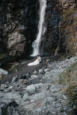Gürültü efekti, seçici odak, manken çekimi: Şelale arka planında çok güzel uzun bir elbise ve damat giymiş çekici bir gelin.