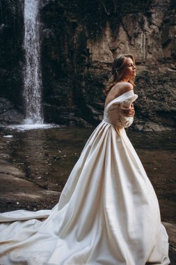 Beyaz elbiseli, sırtı ve omuzları çıplak, güzel bir gelinin zarif ve zarif portresi şelalenin yanındaki fotoğraf için poz veriyor.