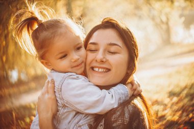 Genç, mutlu ve gülümseyen bir anne kucağında küçük kızıyla sonbahar parkında bir hafta sonu yürüyüşü yapıyor. seçici odak, gürültü etkisi,