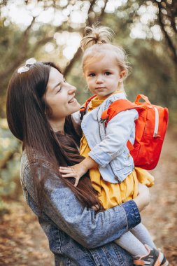 Genç, mutlu ve gülümseyen bir anne kucağında küçük kızıyla sonbahar parkında bir hafta sonu yürüyüşü yapıyor. seçici odak, gürültü etkisi,