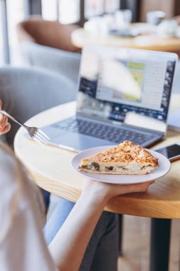 Elinde bir parça et ve mantarlı yulaf lapası olan bir kız bir kafede yemek yerken bilgisayar üzerinde çalışıyor.