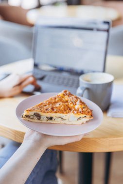 Elinde bir parça et ve mantarlı yulaf lapası olan bir kız bir kafede yemek yerken bilgisayar üzerinde çalışıyor.