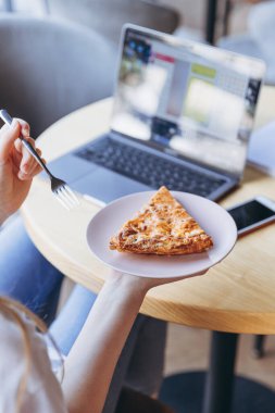 Elinde bir parça et ve mantarlı yulaf lapası olan bir kız bir kafede yemek yerken bilgisayar üzerinde çalışıyor.