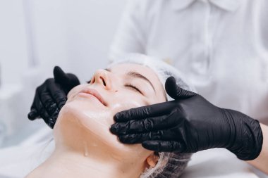 Yüzünde spa ameliyatı olan genç bir kadın. Spa masasına uzanmış bir kadın ve kozmetik maske takan bir kozmetik uzmanı. Metnin için boşluk kopyala