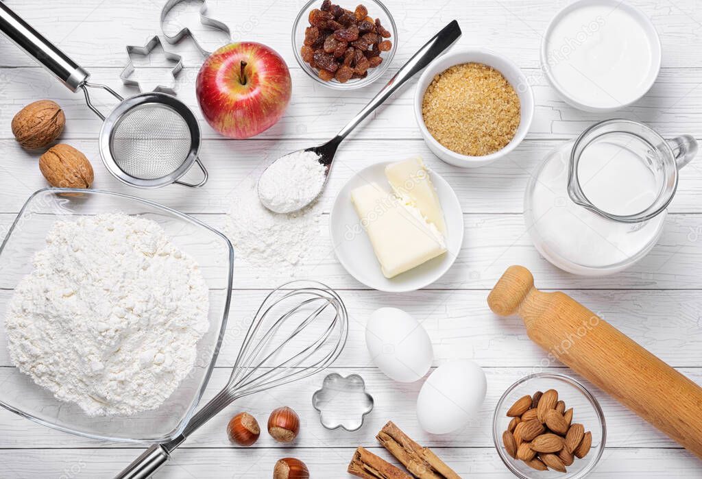 Baking ingredients and tools: flour, eggs, milk, butter, brown sugar, cinnamon, almonds, nuts, yogurt, raisins, walnut and apple with eggbeater and rolling pin on white wooden background, top view.