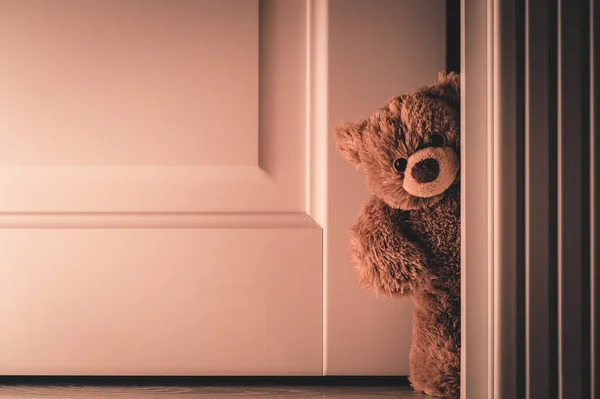 Teddy Bear with red bow peeking out from behind the door. Plush toy looks out of the corner of a wooden door from the bedroom. Flirty flirting in the bedroom.