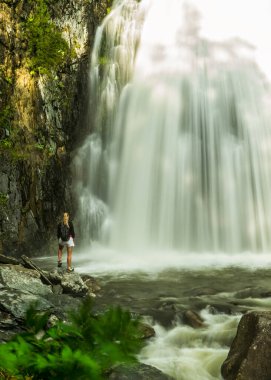 Dökülen kaynak suyunun altındaki bir kayanın üzerinde duran güzel genç bir kadın tropik çağlayan bir şelalenin tadını çıkarıyor. Doğa gezisi, yürüyüş macerası, aile yaz tatili eğlencesi.
