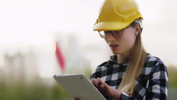 Νεαρή γυναίκα, μηχανικός με κίτρινο σκληρό καπέλο εργάζεται σε tablet. Πορτρέτο, αργή κίνηση — Αρχείο Βίντεο