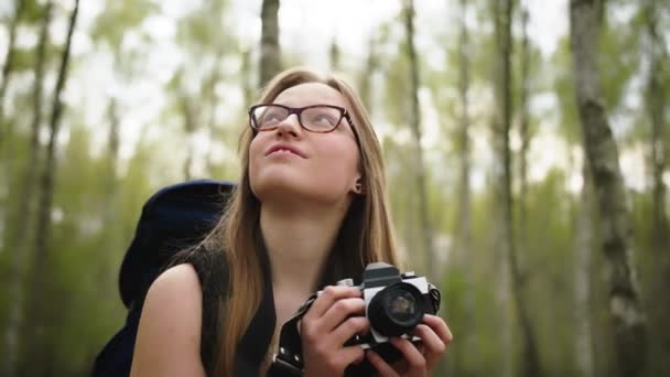 Giovane viaggiatrice caucasica con zaino e macchina fotografica vintage circondata da alberi. Ritratto rallentato — Video Stock