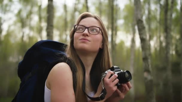 Expresión facial de una joven viajera caucásica con mochila y cámara vintage rodeada de árboles. Retrato de cámara lenta — Vídeo de stock