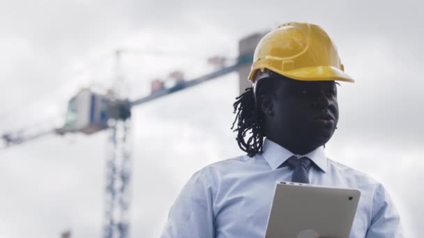 Retrato de um homem africano com chapéu duro usando tablet no canteiro de obras e observando a obra — Vídeo de Stock