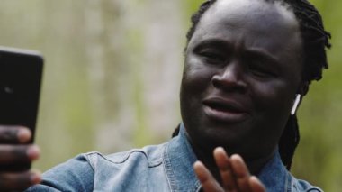 Video görüşmesi için akıllı telefonu kullanan Afrikalı bir adam. Ormandan ailesine el sallıyor. Yavaş çekim. Yakın çekim.