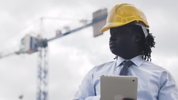 Retrato de um homem africano com chapéu duro usando tablet no canteiro de obras e observando a obra — Vídeo de Stock