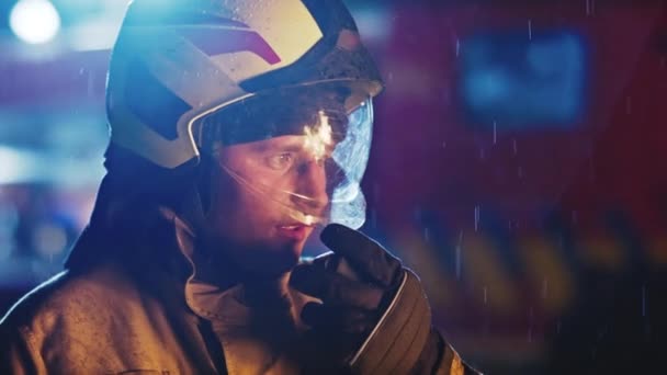 Retrato de un bombero hablando en el walkie talkie y el sitio durante la lluvia. Reflexión de fuego en el casco. Movimiento lento — Vídeos de Stock