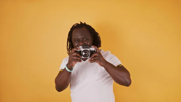 Portret van een Afrikaanse man met gevlochten horen en wit shirt met een vintage camera. Geïsoleerd op de oranje achtergrond — Stockfoto