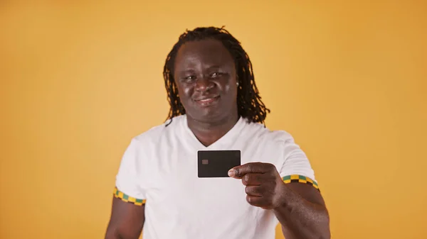 Beyazlı mutlu Afrikalı adam turuncu arka planda kredi kartını kameraya gösteriyor. — Stok fotoğraf