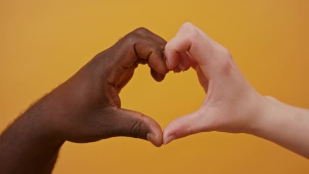 Manos blancas y negras formando forma de corazón juntas aisladas sobre el fondo naranja. De cerca. — Vídeos de Stock