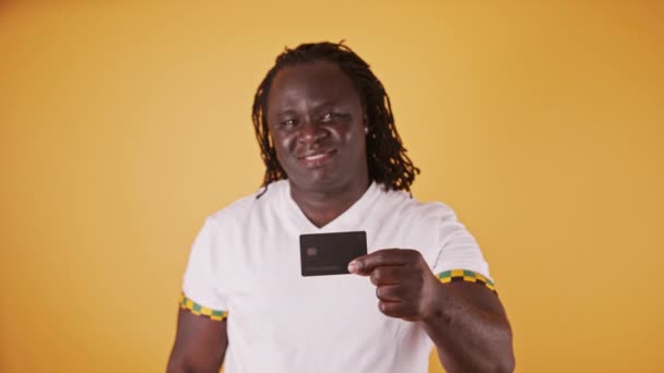 Щасливий африканський чоловік у білому, показуючи кредитну картку на камеру на помаранчевому фоні — стокове відео