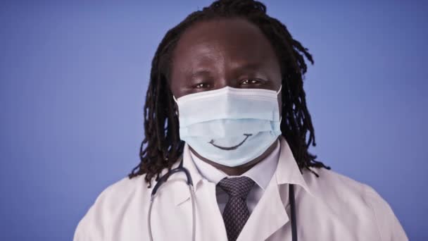 Retrato de um médico africano em máscara facial com sorriso isolado no fundo do estúdio azul — Vídeo de Stock