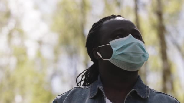 Retrato de un hombre africano con máscara médica y auriculares inalámbricos en la naturaleza — Vídeos de Stock