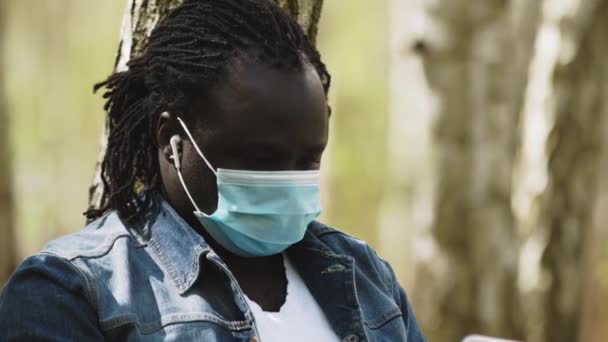 Koncept vzdálené práce. Afričan s lékařskou maskou pomocí notebooku v parku — Stock video