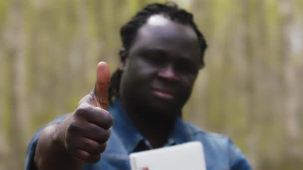 Homem africano com um tablet segurando um polegar no parque. Foco da cremalheira — Vídeo de Stock