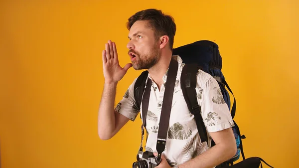 Young man traveler with backpack speaking loud with hands on the side of his face isolated on the orange background — Stock Photo, Image