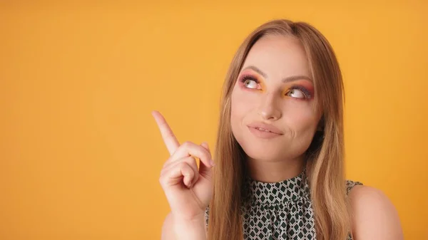 Porträt einer jungen modischen Frau mit Make-up, die mit dem Finger auf den leeren Raum zeigt, isoliert auf orangefarbenem Hintergrund. Kopierraum — Stockfoto