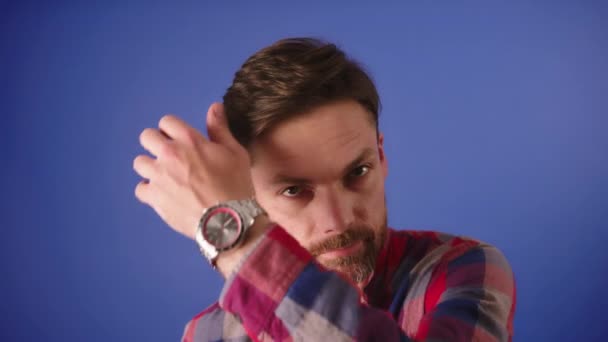 Mladý flirtující běloch s vousy si upravuje vlasy. Portrét izolovaný na modrém pozadí — Stock video