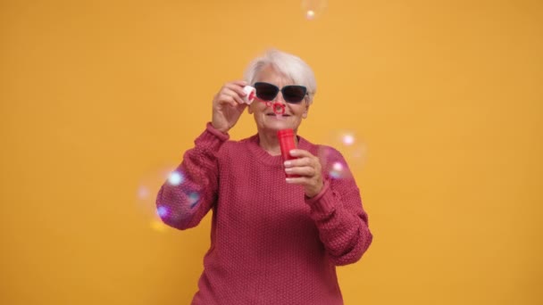 선글라스를 끼고 긴 산뜻 한 스웨터를 입은 유행하는 할머니가 비누 거품을 불고 있다. 고립 됨 — 비디오