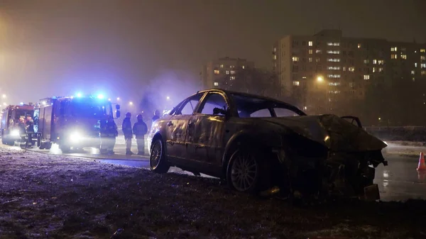 Winter car accident, burned car with fire engine and buildings in the background